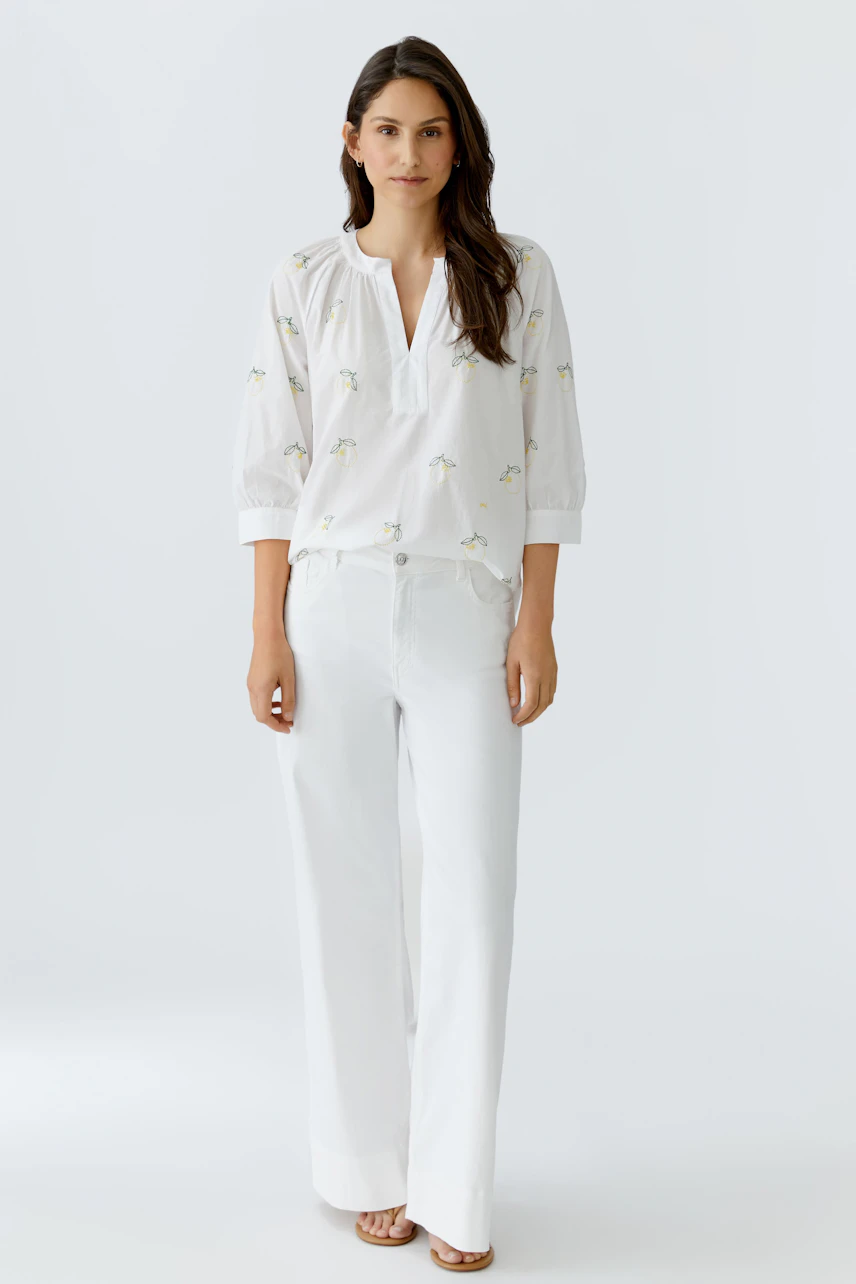 Lässige Damen-Tunika: Frau in weißer Tunika mit dezentem Muster und weiter Hose steht entspannt.