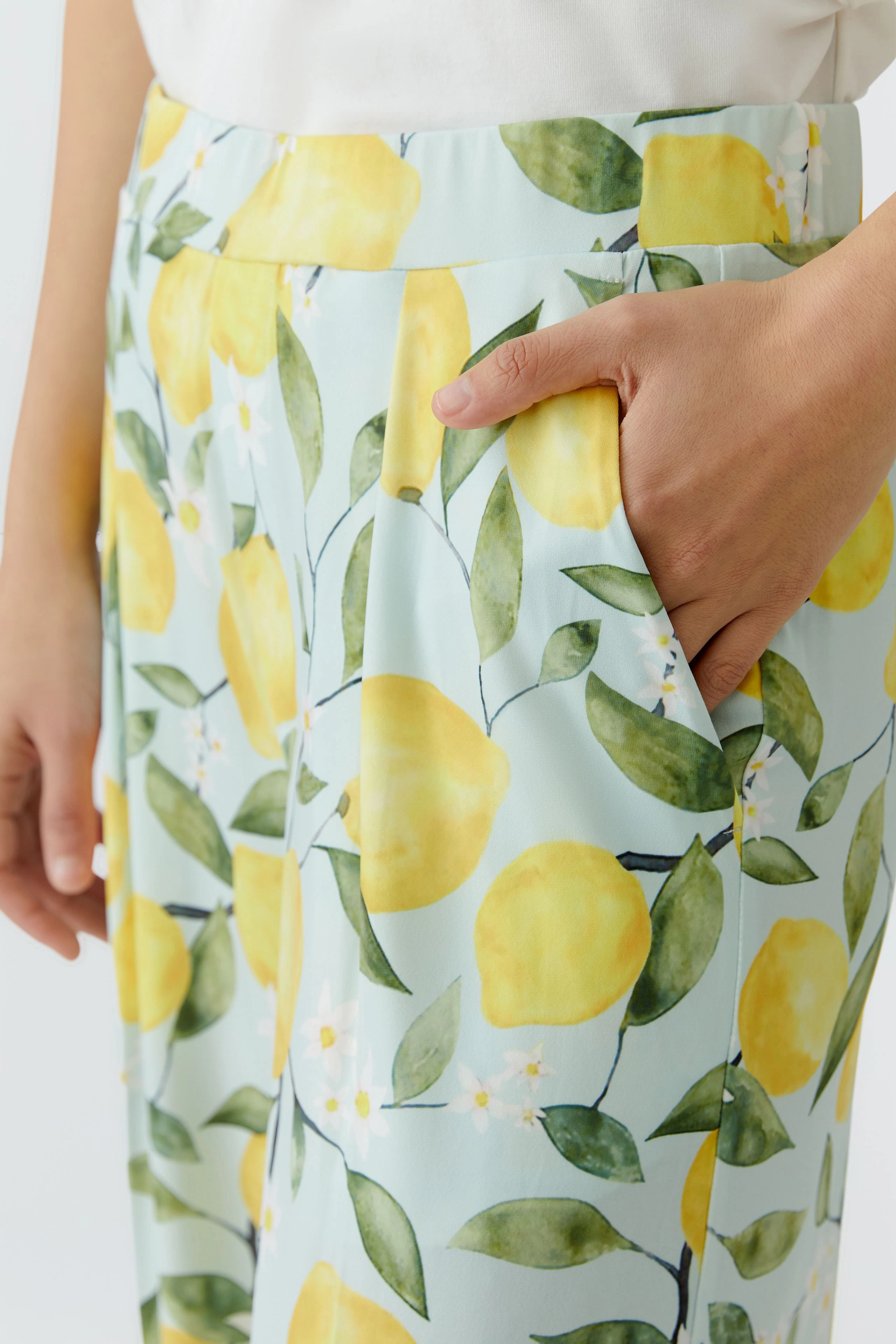 Woman in lemon-patterned pants with a white top, hand in pocket, showcasing vibrant summer style.