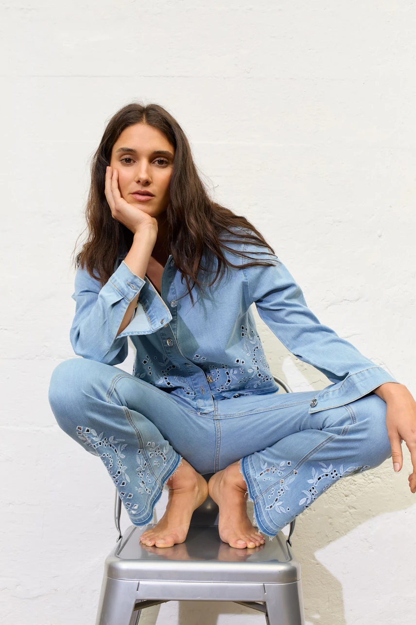 Casual denim jacket: Woman in light blue jeans jacket with floral cutouts, sitting confidently on a chair.
