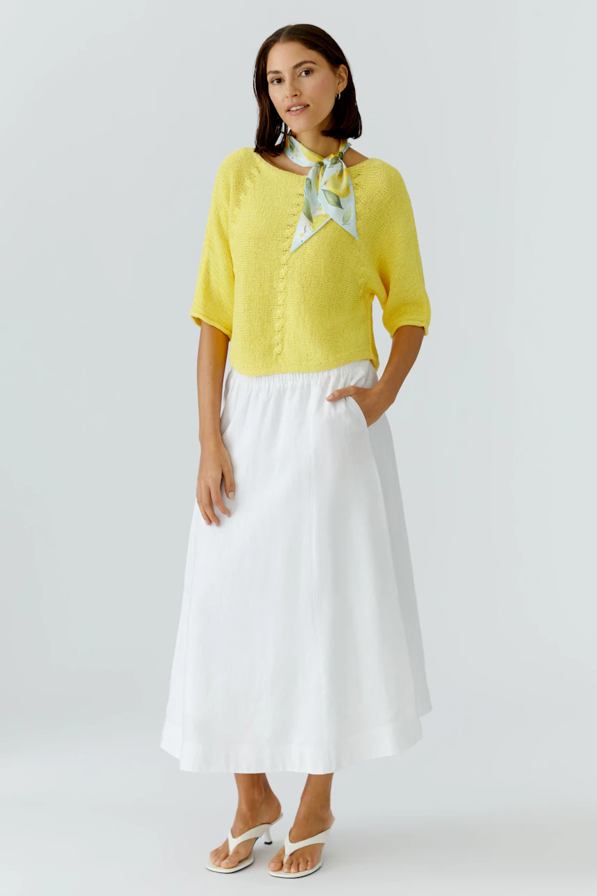 Woman in yellow pullover with a colorful scarf, wearing a white skirt and sandals.