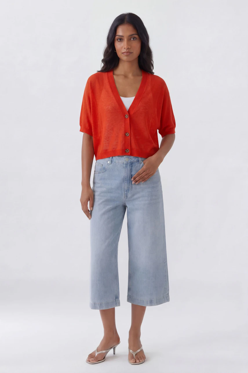 Casual cardigan: Woman in vibrant red knit cardigan, white top, and light blue wide-leg jeans, standing confidently.