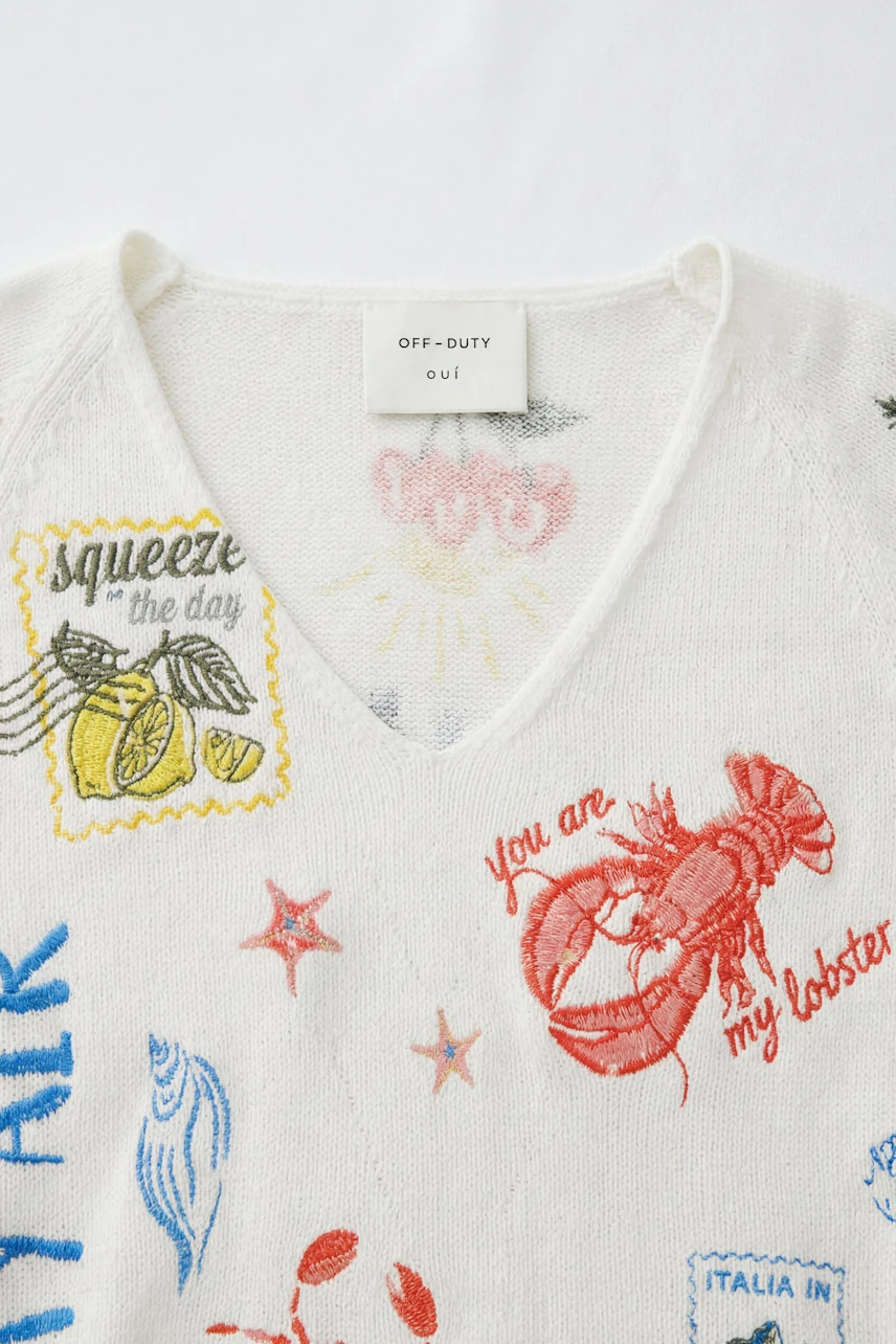 White pullover with colorful embroidery, including a lobster and lemons, on a flat surface.