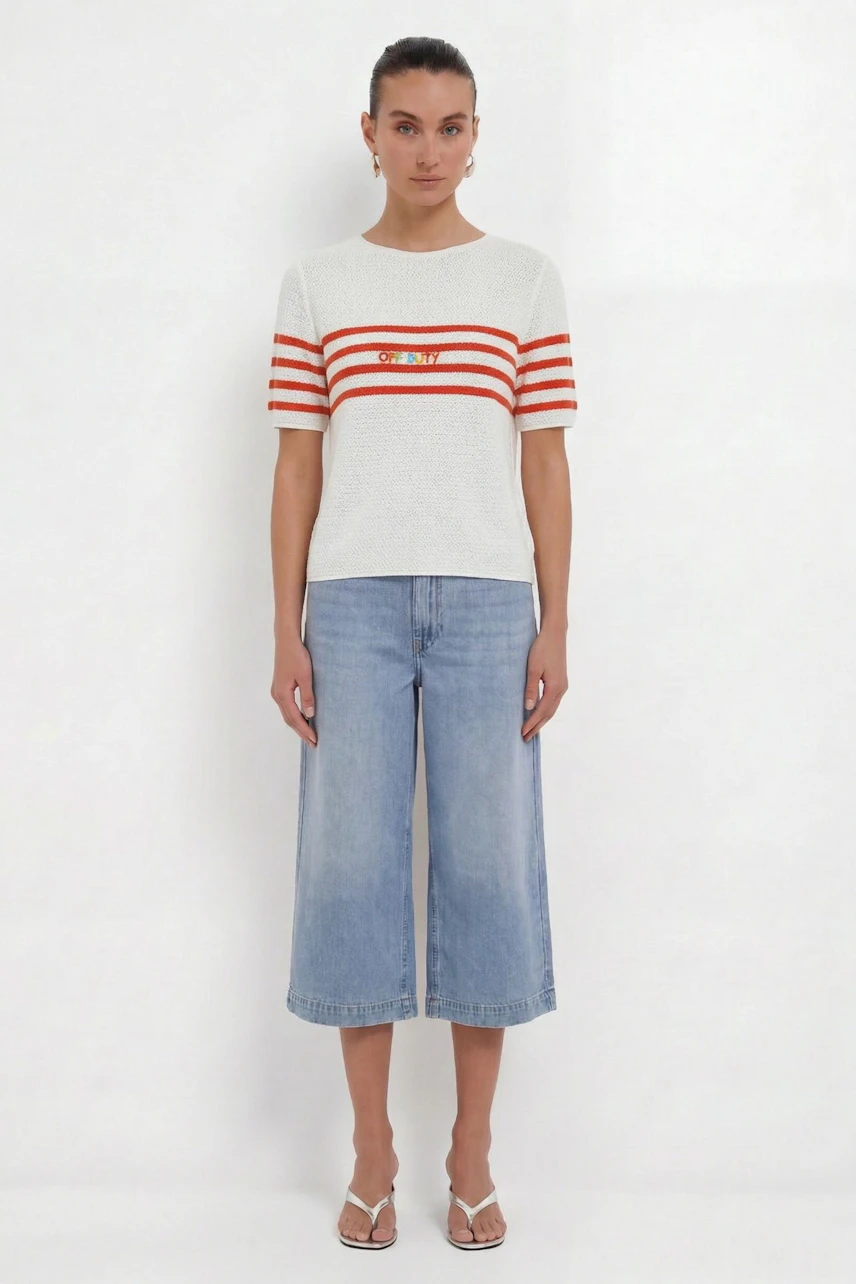 Frau im lässigen Pullover mit roten Streifen und Logo, kombiniert mit Jeans-Culottes und Sandalen.