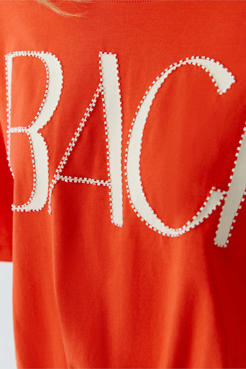 Bright orange T-shirt with bold white letters and studded details, worn by a woman.