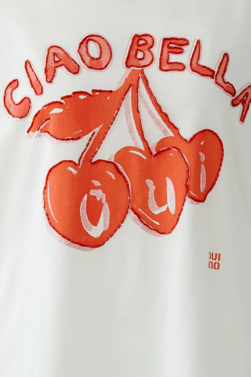 Frau im weißen T-Shirt mit rotem Print "Ciao Bella" und Kirschen, lässiger Look.