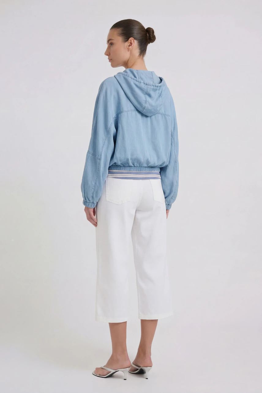 Casual women's jacket: Woman in a light blue hooded jacket with white cropped pants, standing confidently.