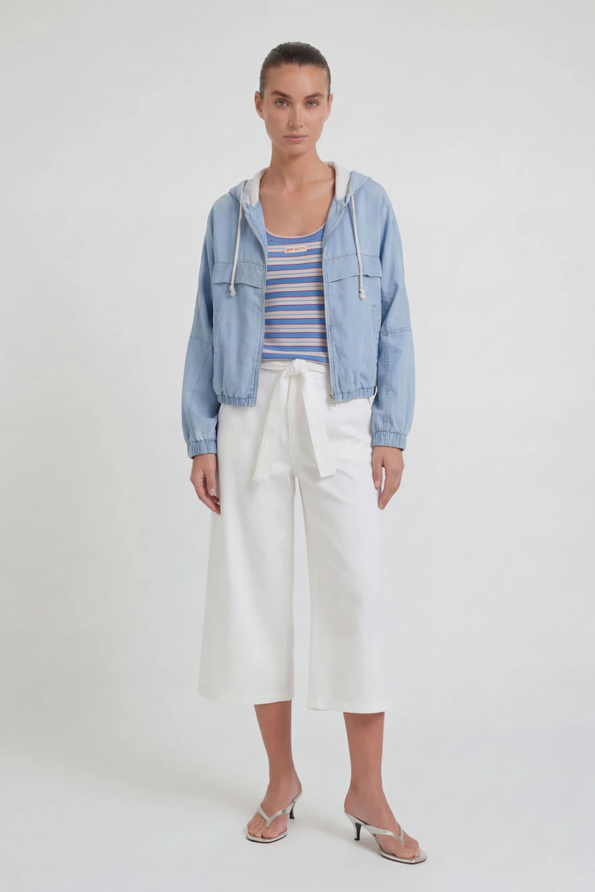 Casual women's jacket: Woman in light blue jacket over striped top and white pants, standing confidently.