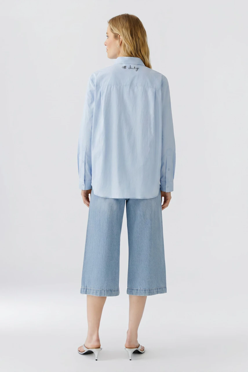 Lässige Damen-Bluse: Frau in hellblauer Bluse und weißen Hosen, von hinten gesehen.