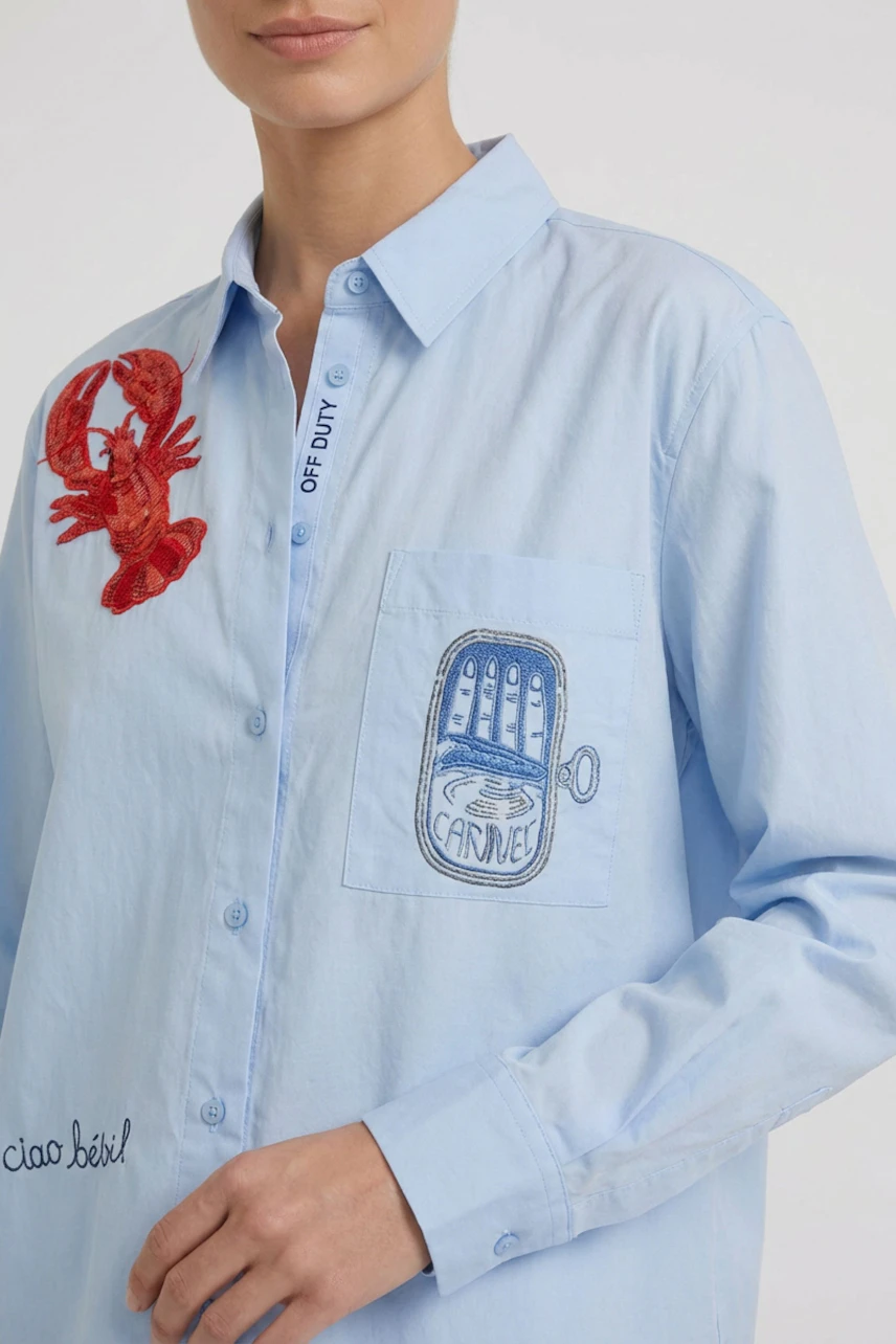 Casual women's blouse: Woman in light blue blouse with red lobster and blue can embroidery, relaxed fit.