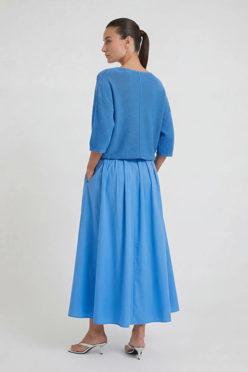 Woman in a blue midi skirt and matching top, standing sideways, hands in pockets, wearing white heels.