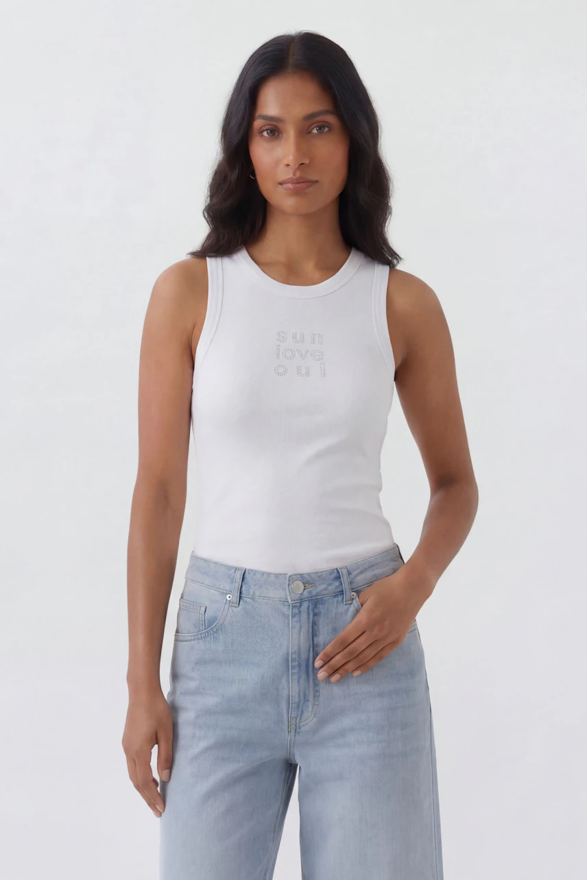 Casual tank top: Woman in white tank top with subtle text, paired with light blue jeans, standing confidently.