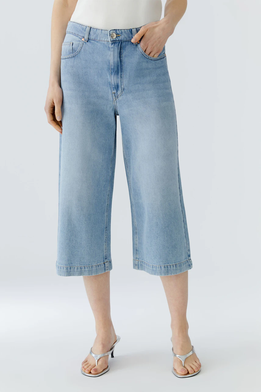 Casual culotte: Woman in light blue denim culotte, white top, and sandals, standing confidently.