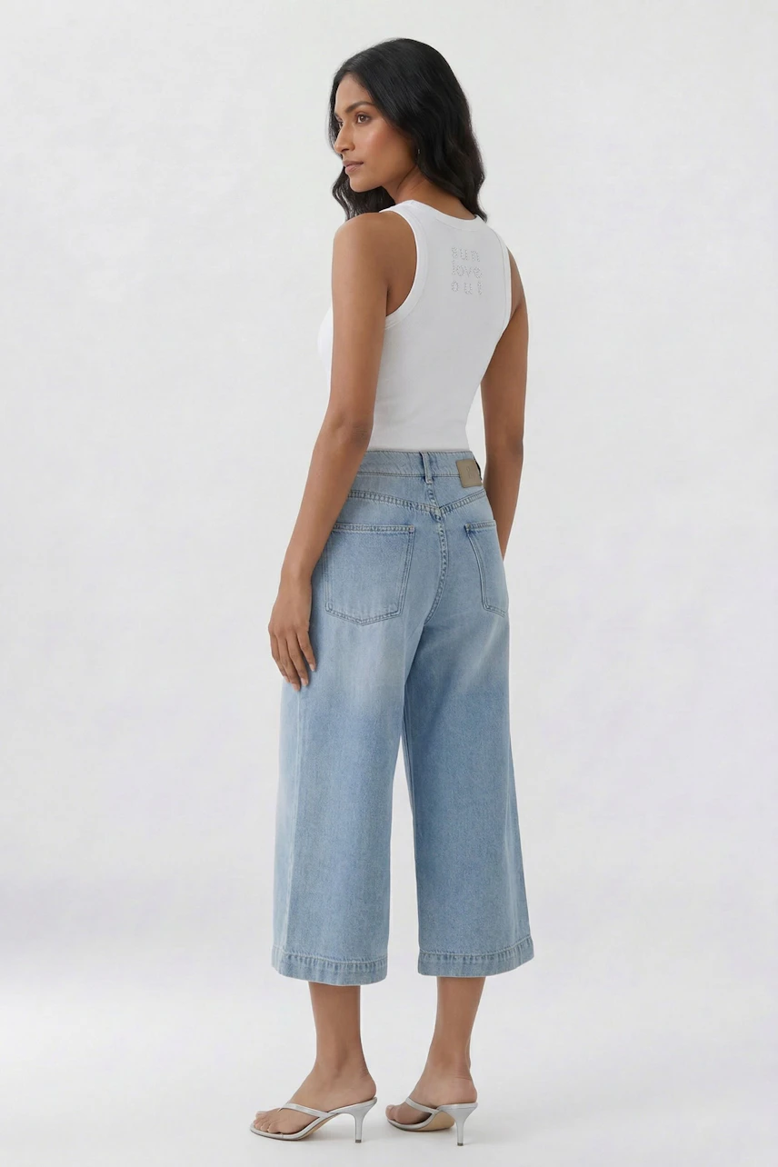 Woman in light blue denim culotte and white tank top, standing sideways, showcasing casual style.