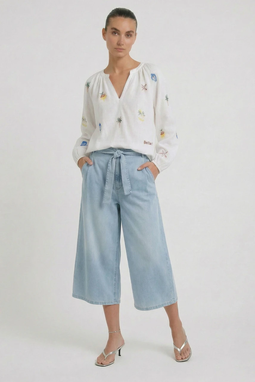 Woman in light blue culotte and embroidered blouse, hands in pockets, wearing sandals.