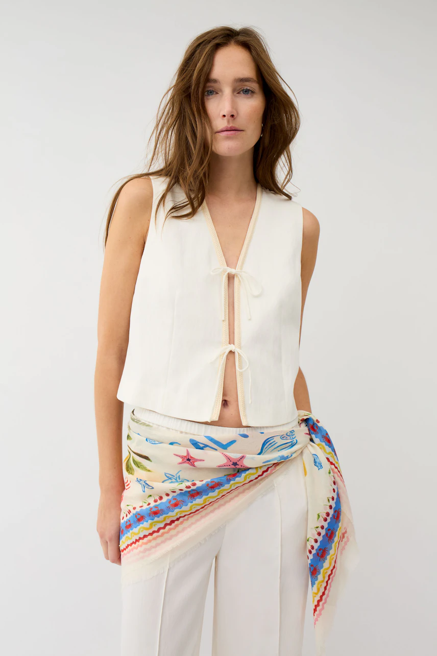 Woman wearing a colorful scarf tied at the waist over white pants and a sleeveless top.