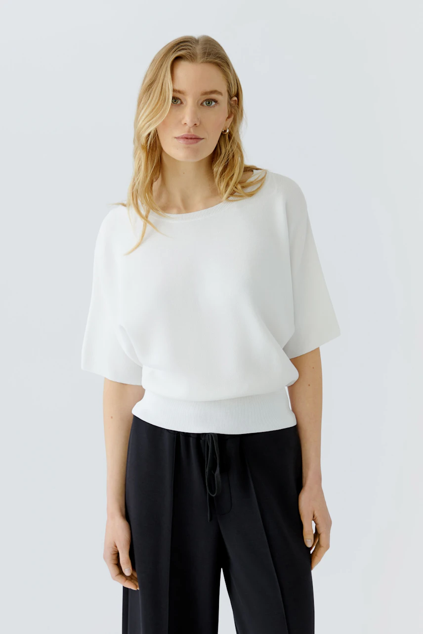 Casual women's pullover: Woman in a white pullover with loose sleeves and black pants, standing confidently.