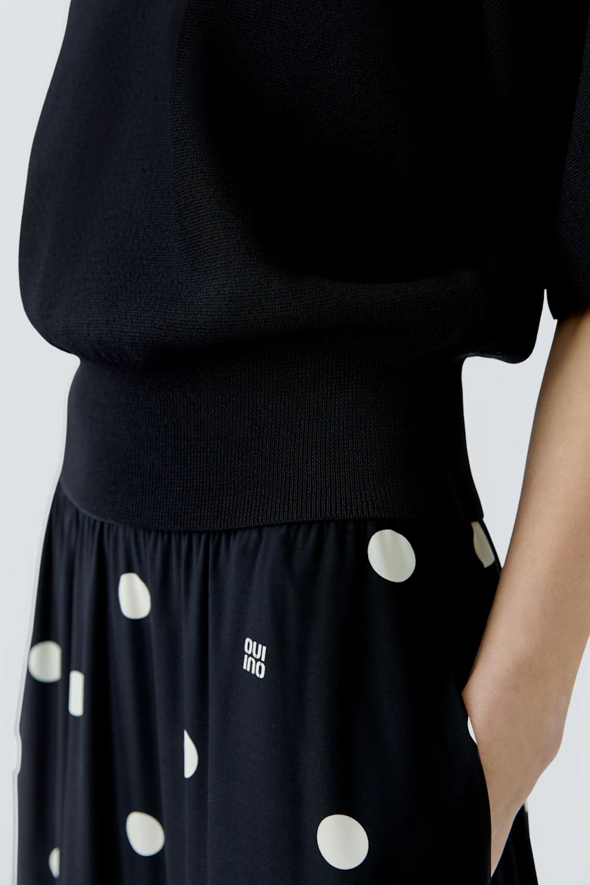 Casual pullover: Woman in a black pullover with a polka dot skirt, hands in pockets, relaxed style.