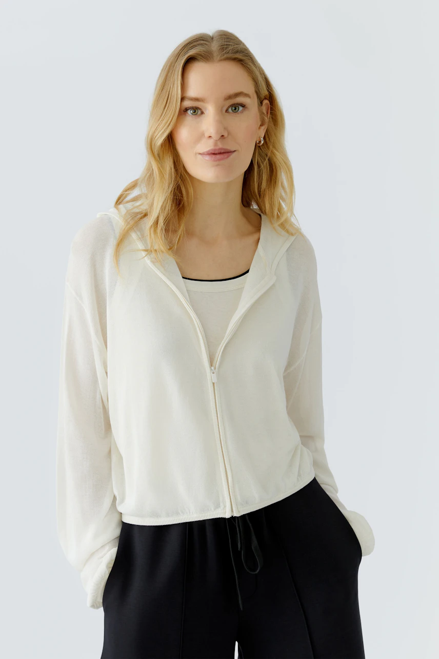 Lässige Strickjacke: Frau in weißer Strickjacke mit Kapuze und schwarzem Top, entspannt und modern.