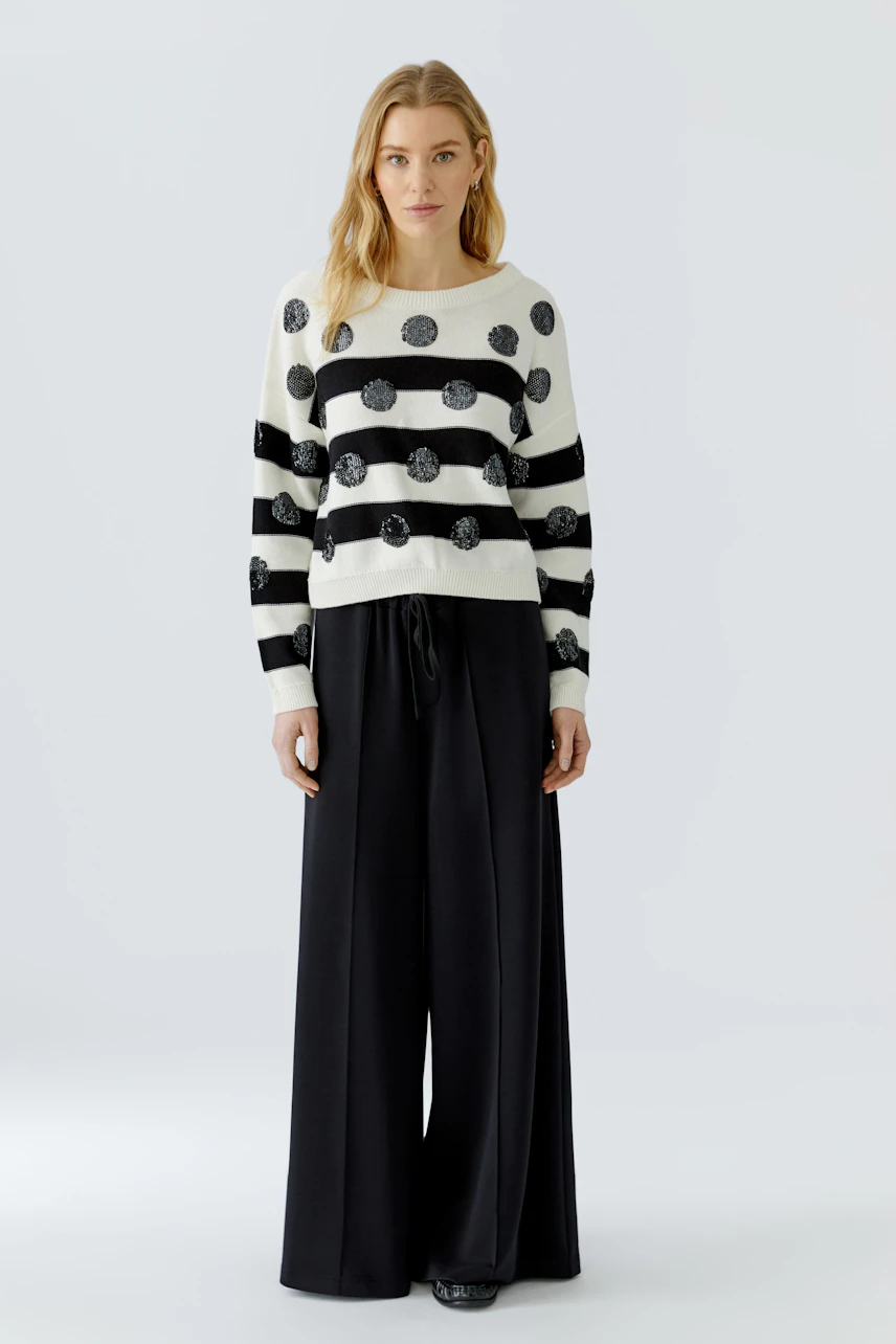 Casual women's pullover: Woman in a white pullover with black stripes and dots, paired with wide black pants.