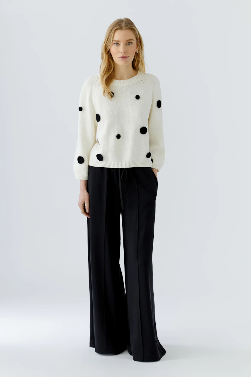 Casual women's pullover: Woman in white pullover with black dots and wide black pants stands confidently.