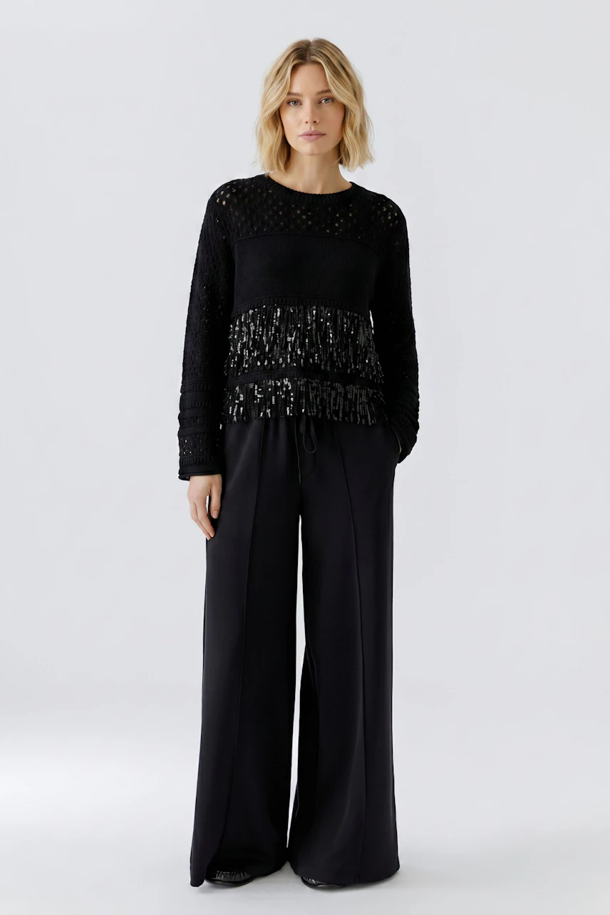 Woman in a black pullover with fringe details, paired with wide-leg pants, standing confidently.