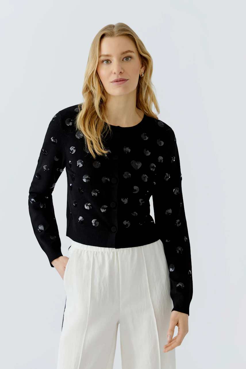 Woman in black cardigan with polka dots, paired with white pants, looking confidently.