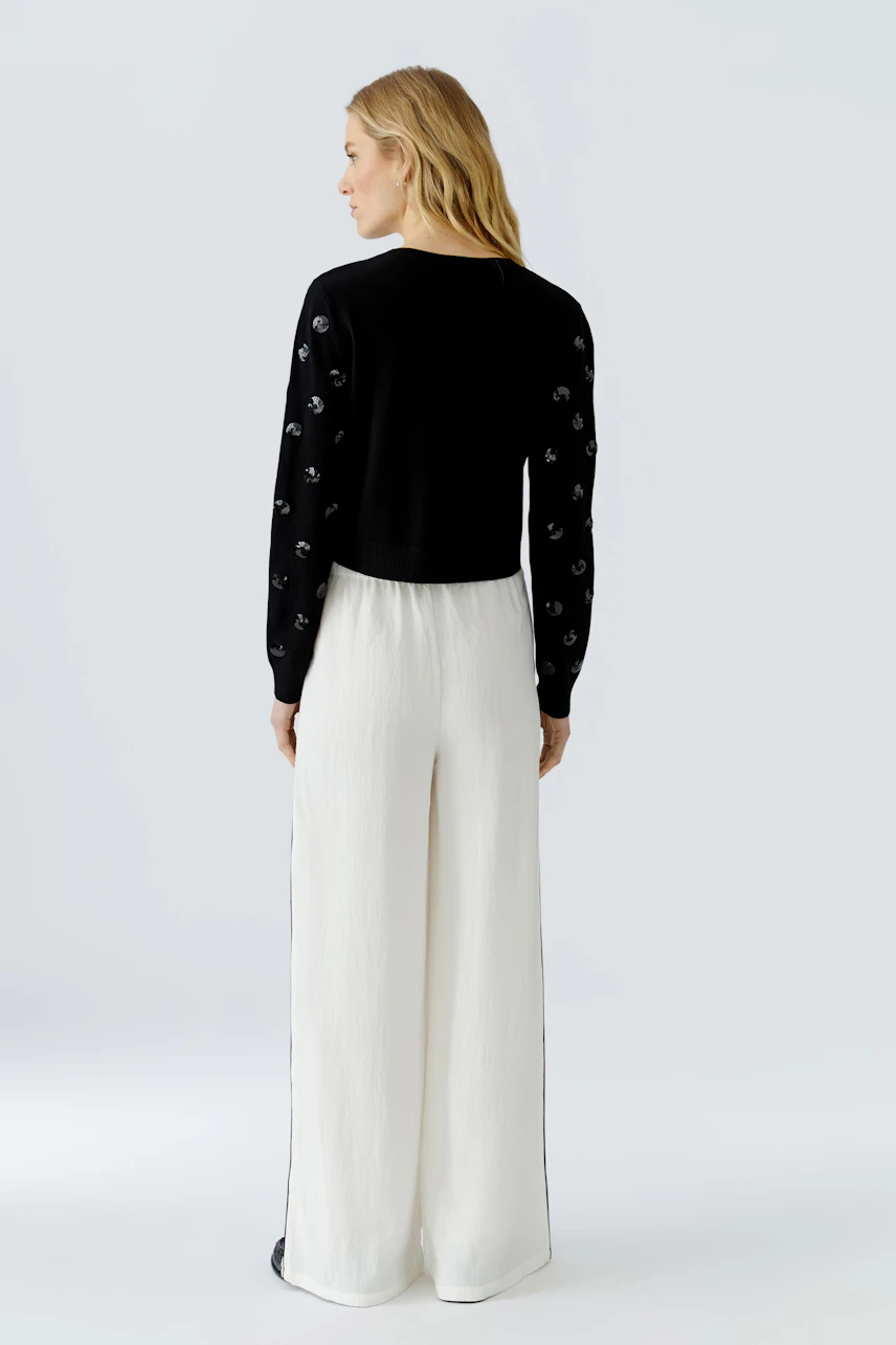 Woman in black cardigan with textured sleeves, paired with white wide-leg pants, looking to the side.