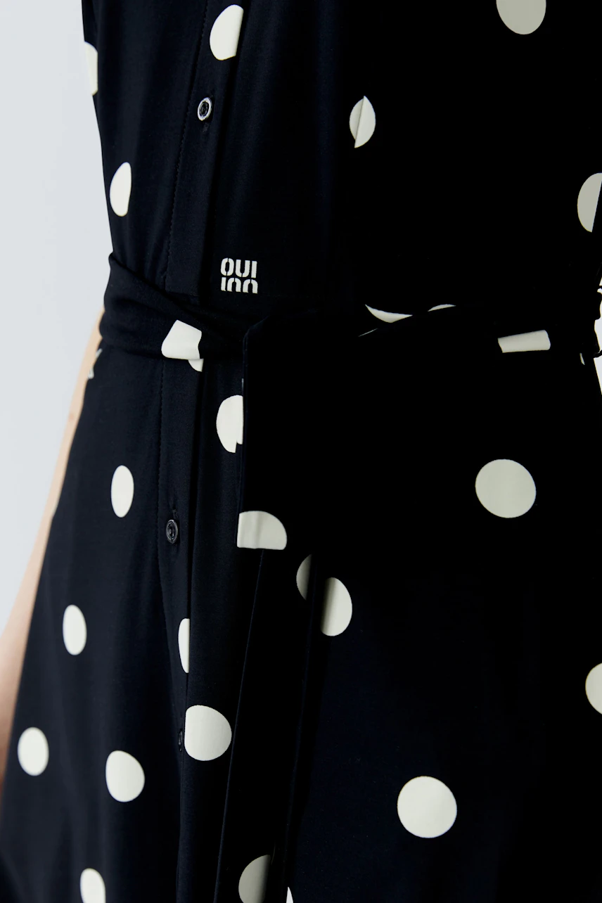 Schwarzes Kleid mit weißen Punkten: Frau trägt ein gepunktetes Kleid mit Gürtel, schlicht und stilvoll.