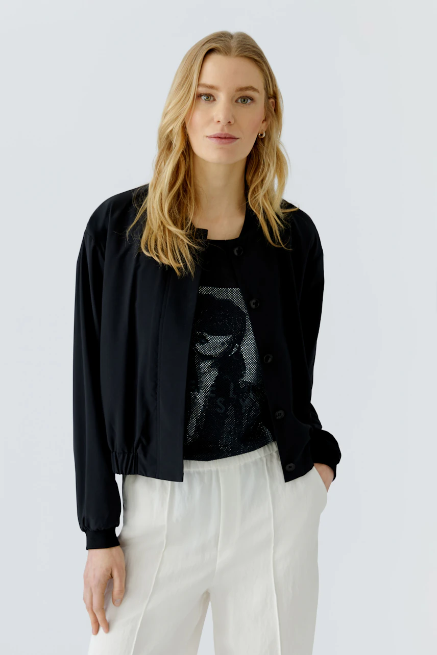 Casual blouson: Woman in a black blouson over a graphic tee, paired with white pants, exudes confidence.