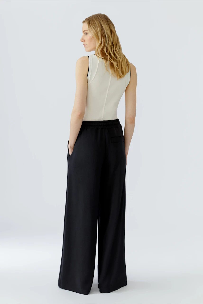Frau in schwarzer Marlenehose und weißem Top, seitlich stehend, zeigt lässigen Look.
