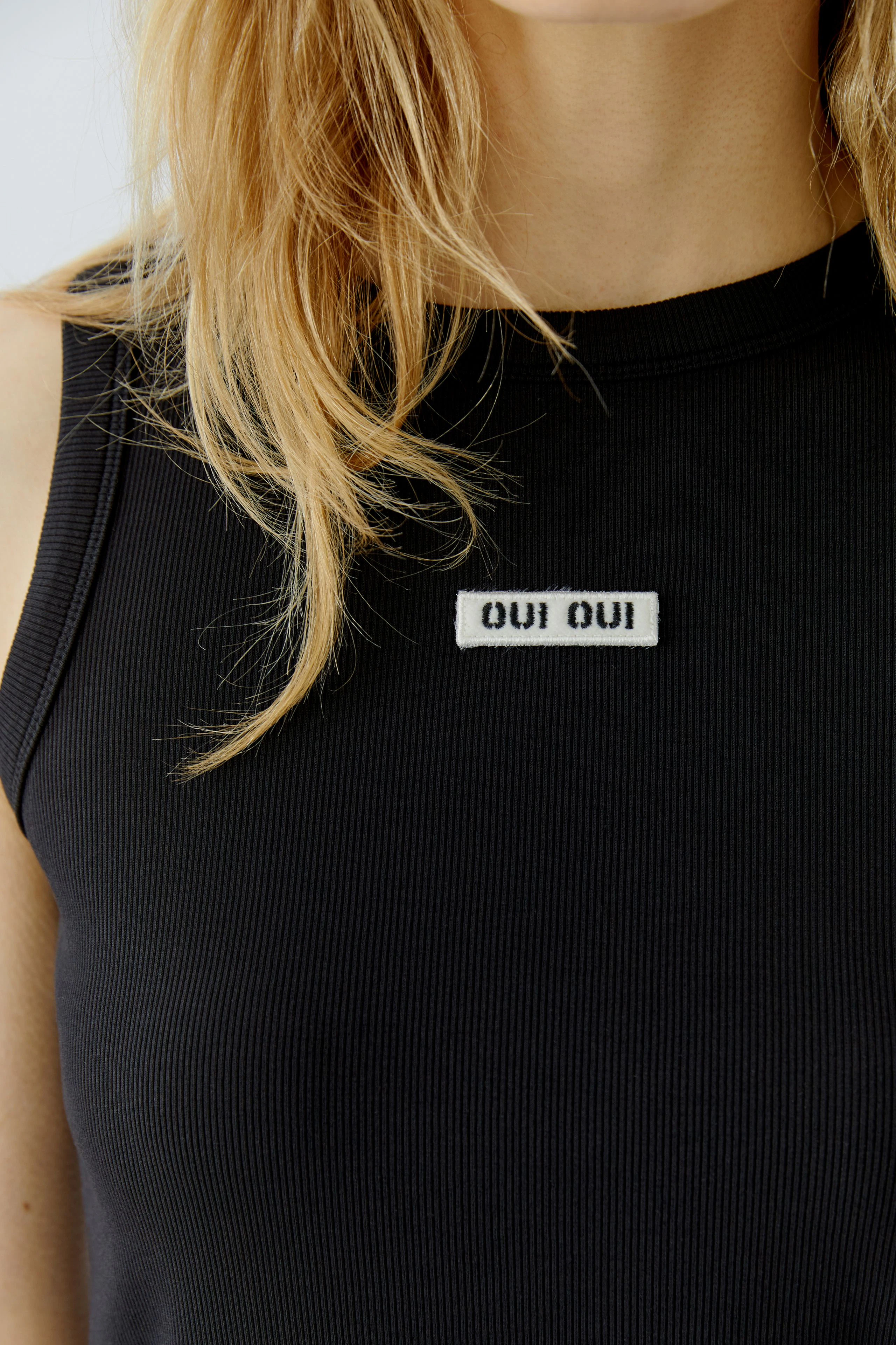Casual tank top: Woman in black ribbed tank top with "Oui Oui" patch, showcasing a relaxed style.