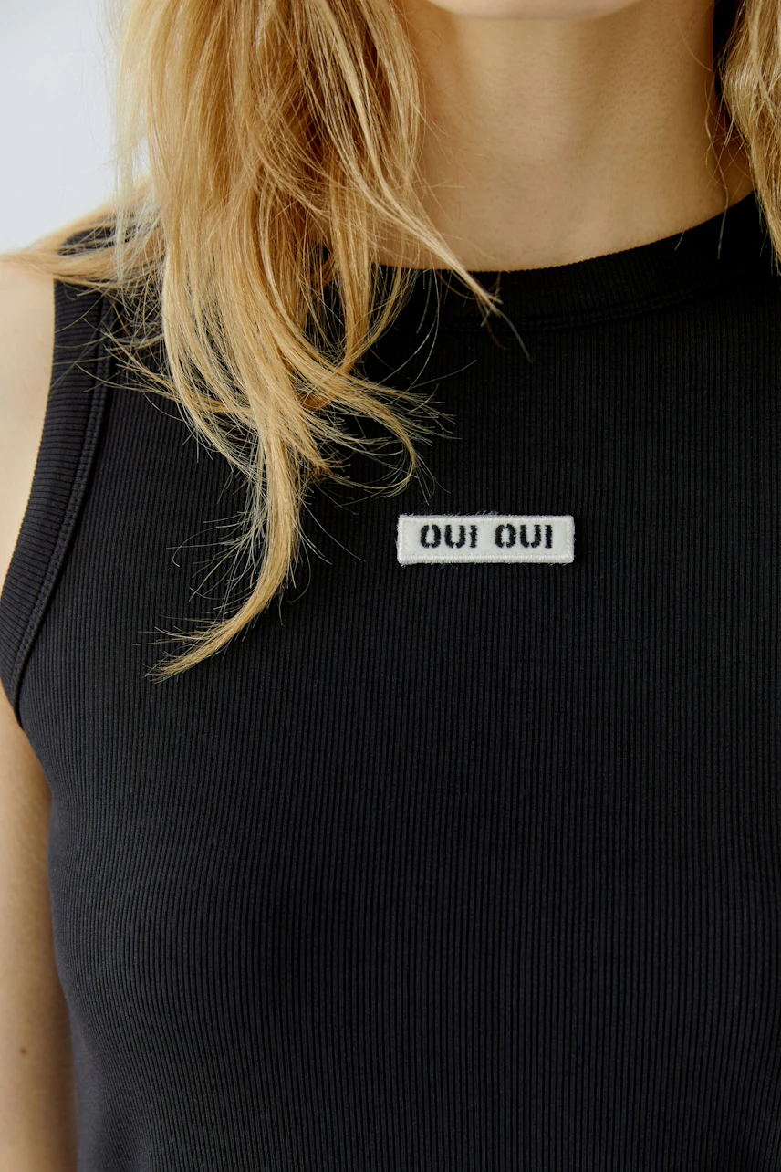 Casual tank top: Woman in black ribbed tank top with "Oui Oui" patch, showcasing a relaxed style.