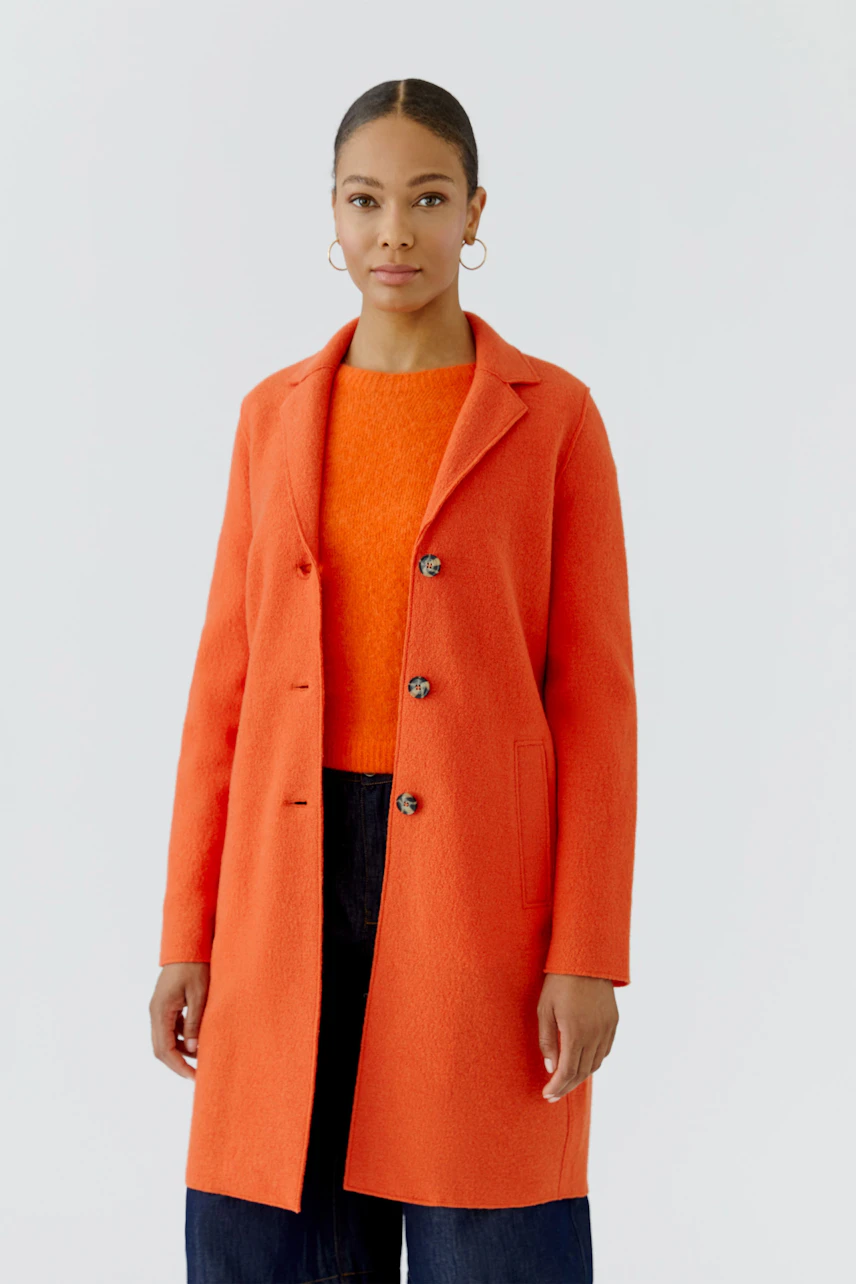 Frau im orangefarbenen Wollmantel, kombiniert mit passendem Pullover und dunkler Hose.
