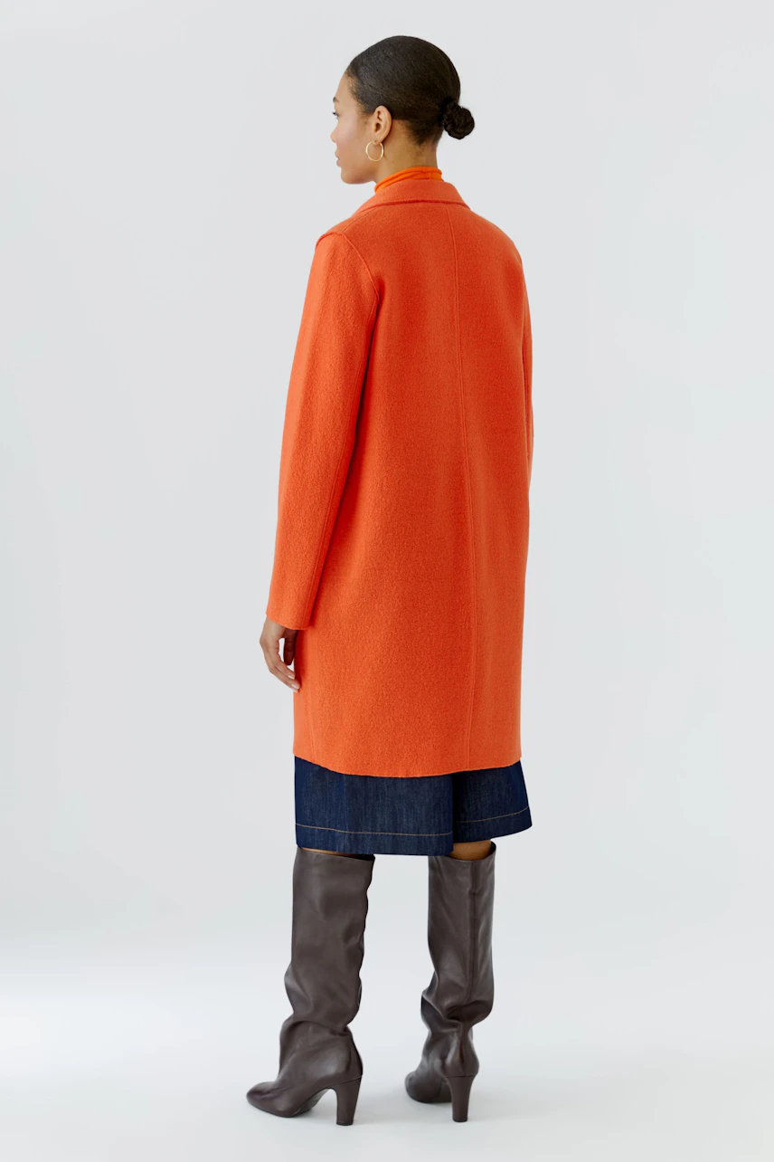 Frau im orangefarbenen Wollmantel, kombiniert mit Jeansrock und braunen Stiefeln, von hinten gesehen.