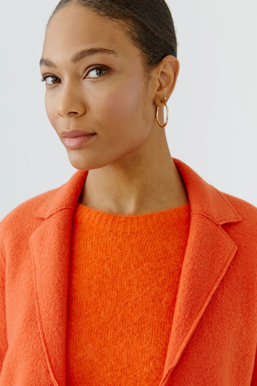 Frau im orangefarbenen Wollmantel: Schlichter Look mit passendem Pullover, dezente Ohrringe.