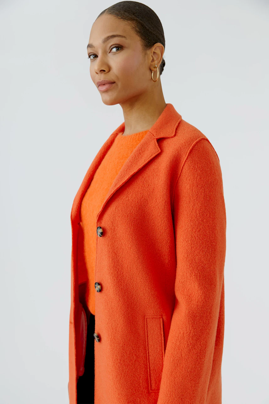 Frau im leuchtend orangefarbenen Wollmantel, schlichtes Design, mit passendem Pullover darunter.