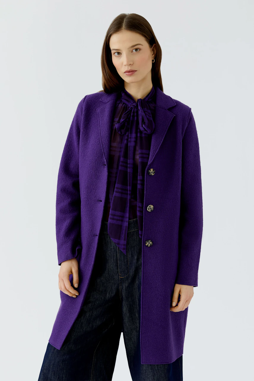 Woman in a purple wool coat over a matching blouse and dark jeans, standing confidently.