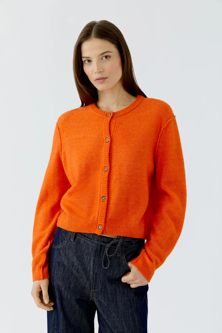 Frau in leuchtend oranger Strickjacke, kombiniert mit dunkler Jeans, schaut selbstbewusst.