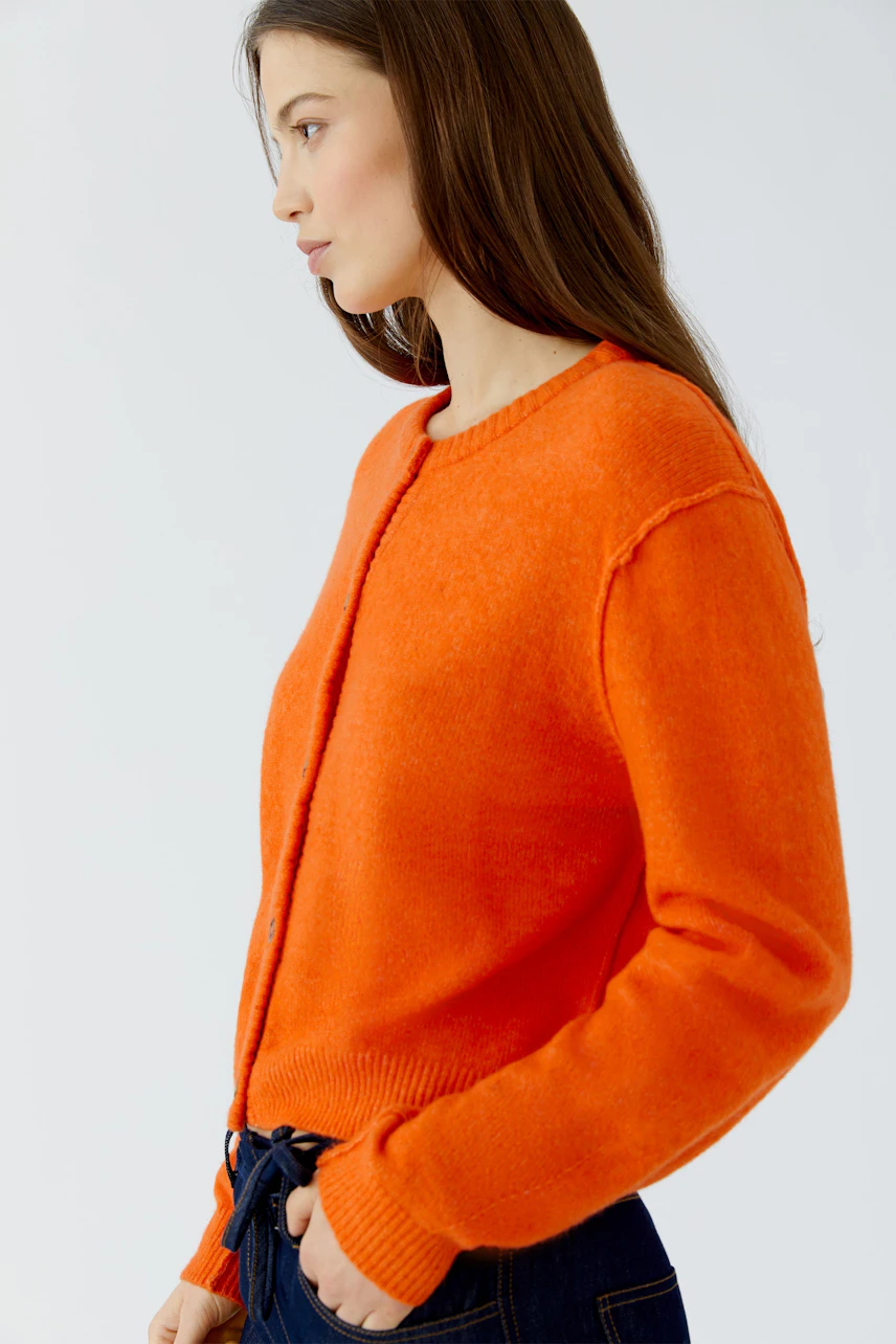 Frau in leuchtend orangefarbener Strickjacke, seitlich betrachtet, kombiniert mit dunkler Jeans.