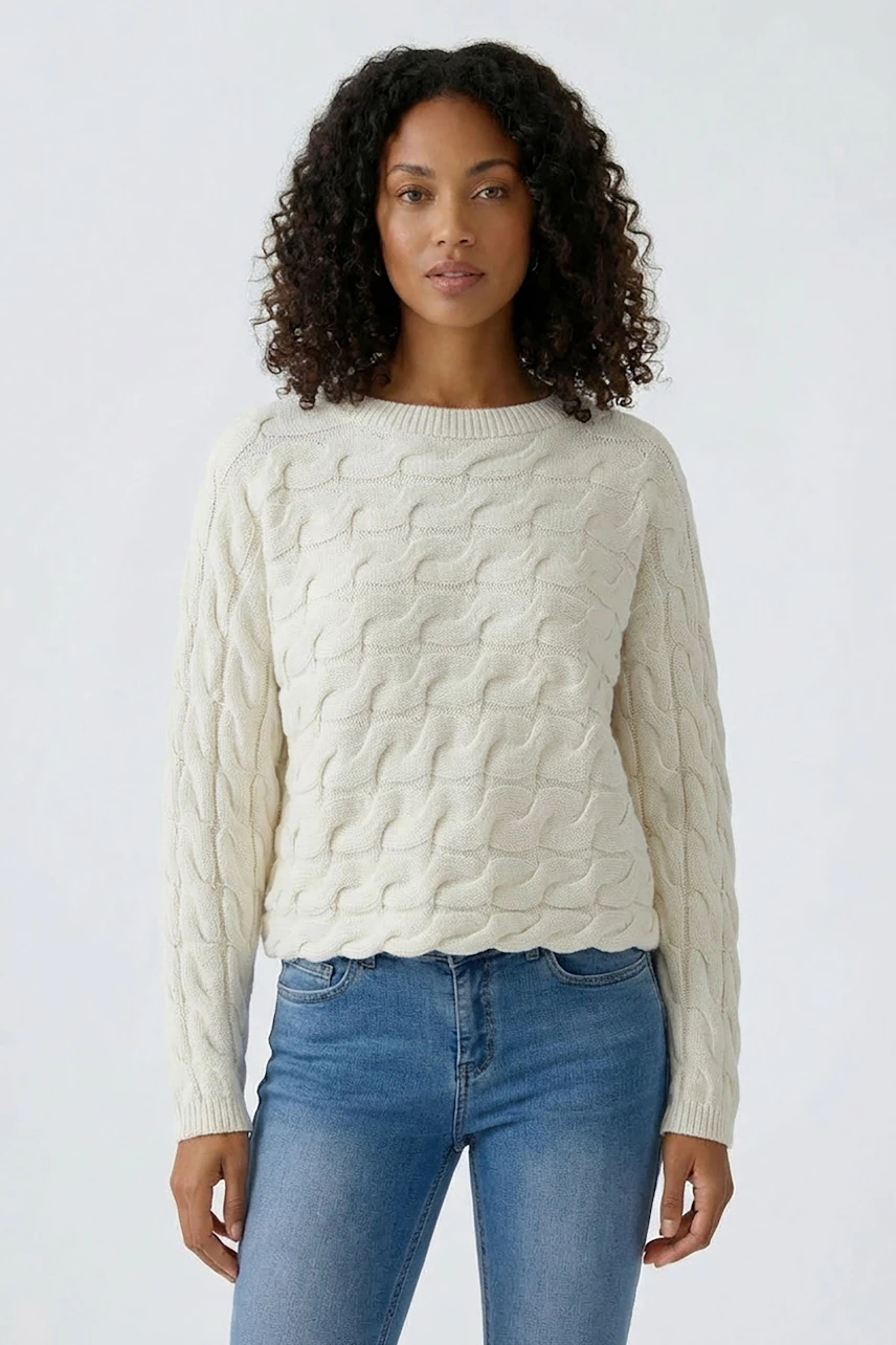 Casual women's pullover: Woman in a cream cable-knit pullover and blue jeans, standing confidently.