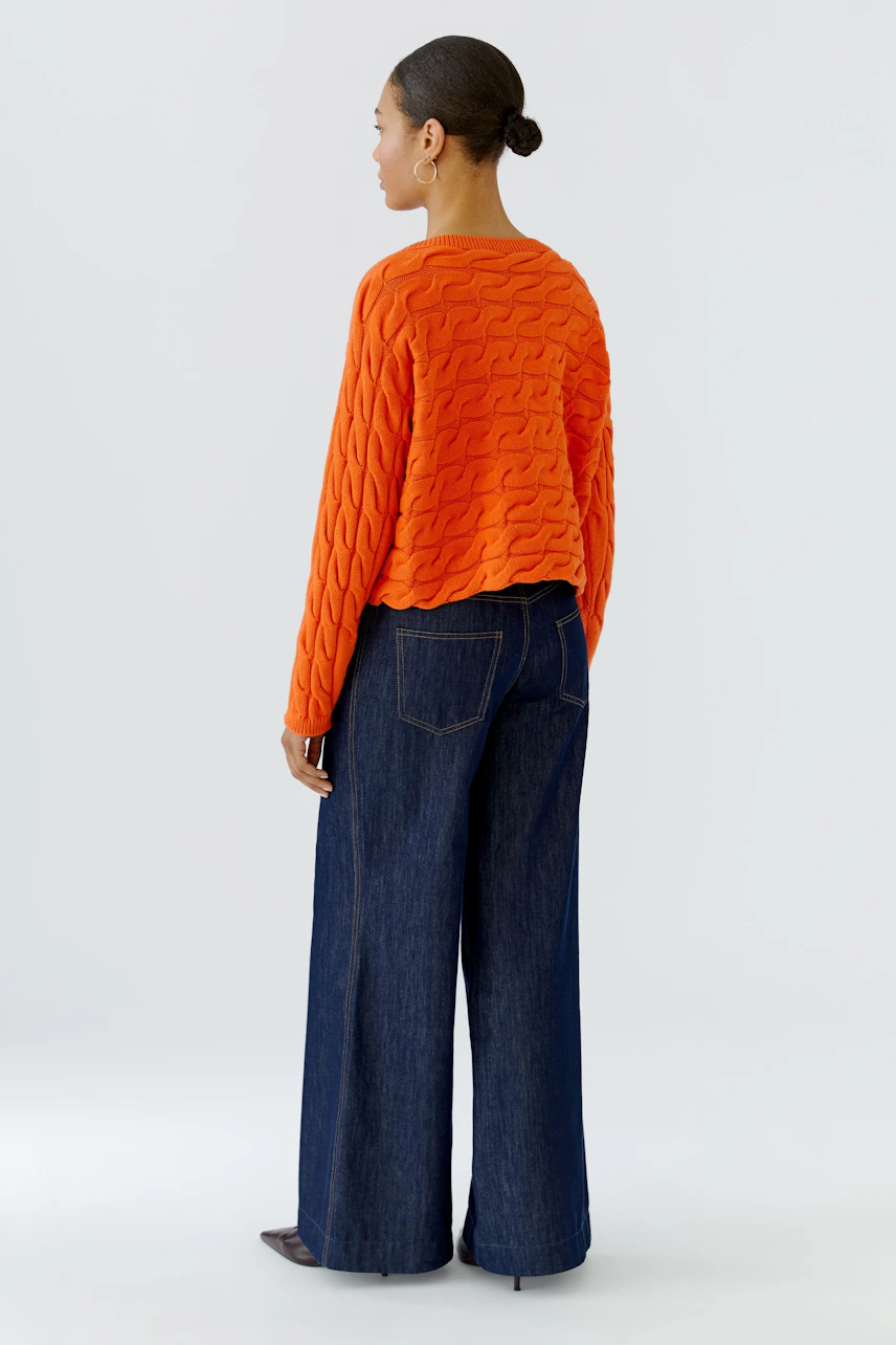 Casual women's pullover: Woman in vibrant orange pullover with textured pattern, paired with wide-leg jeans.