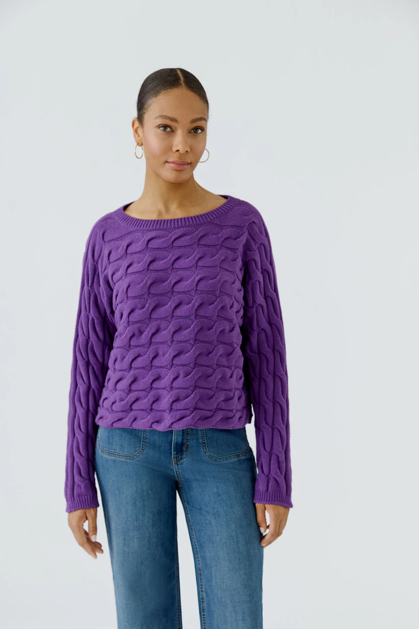 Frau im lila Pullover mit Zopfmuster, kombiniert mit blauer Jeans. Schlicht und modern.