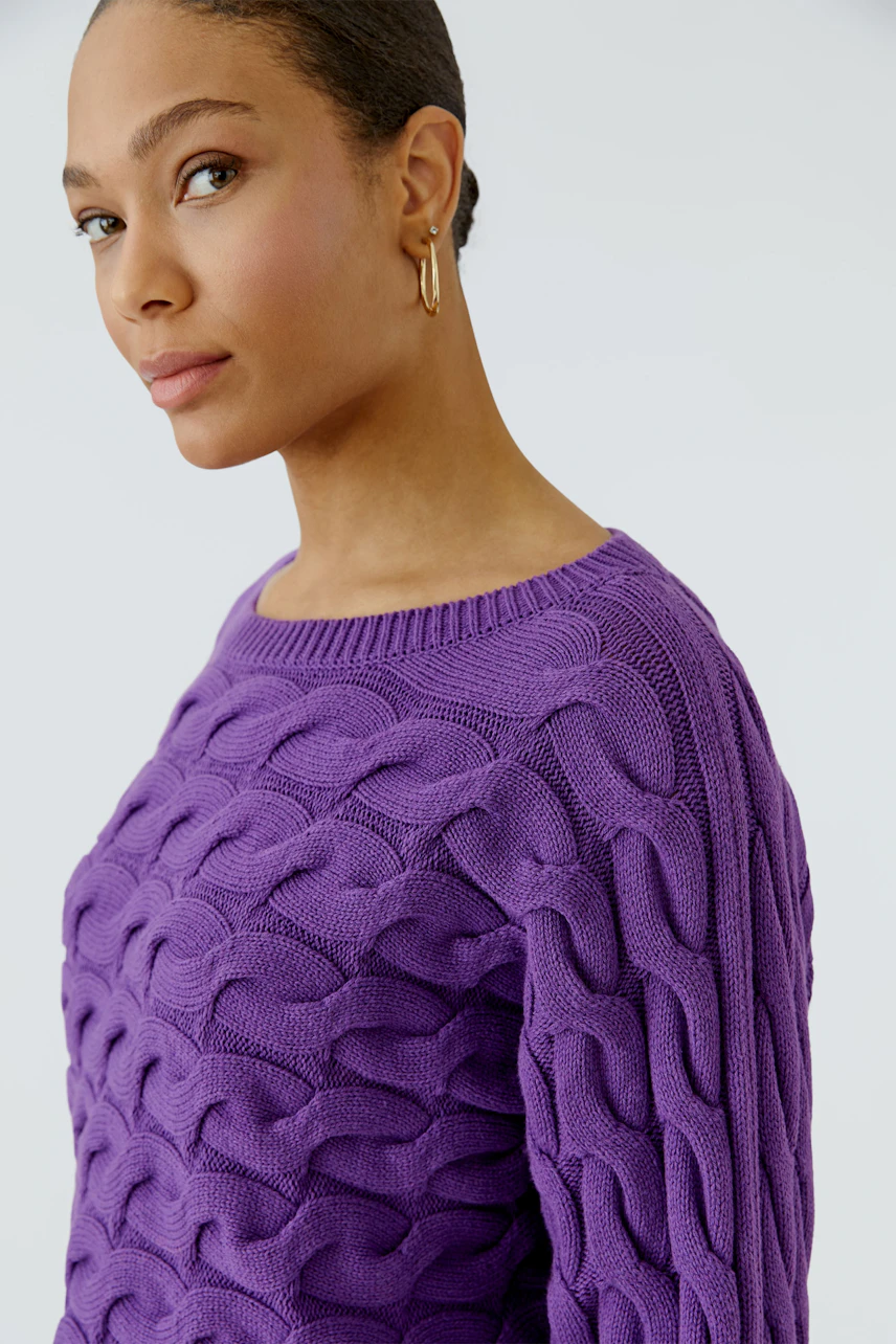 Frau im lila Pullover mit Zopfmuster, schaut zur Seite. Der Pullover hat einen Rundhalsausschnitt.