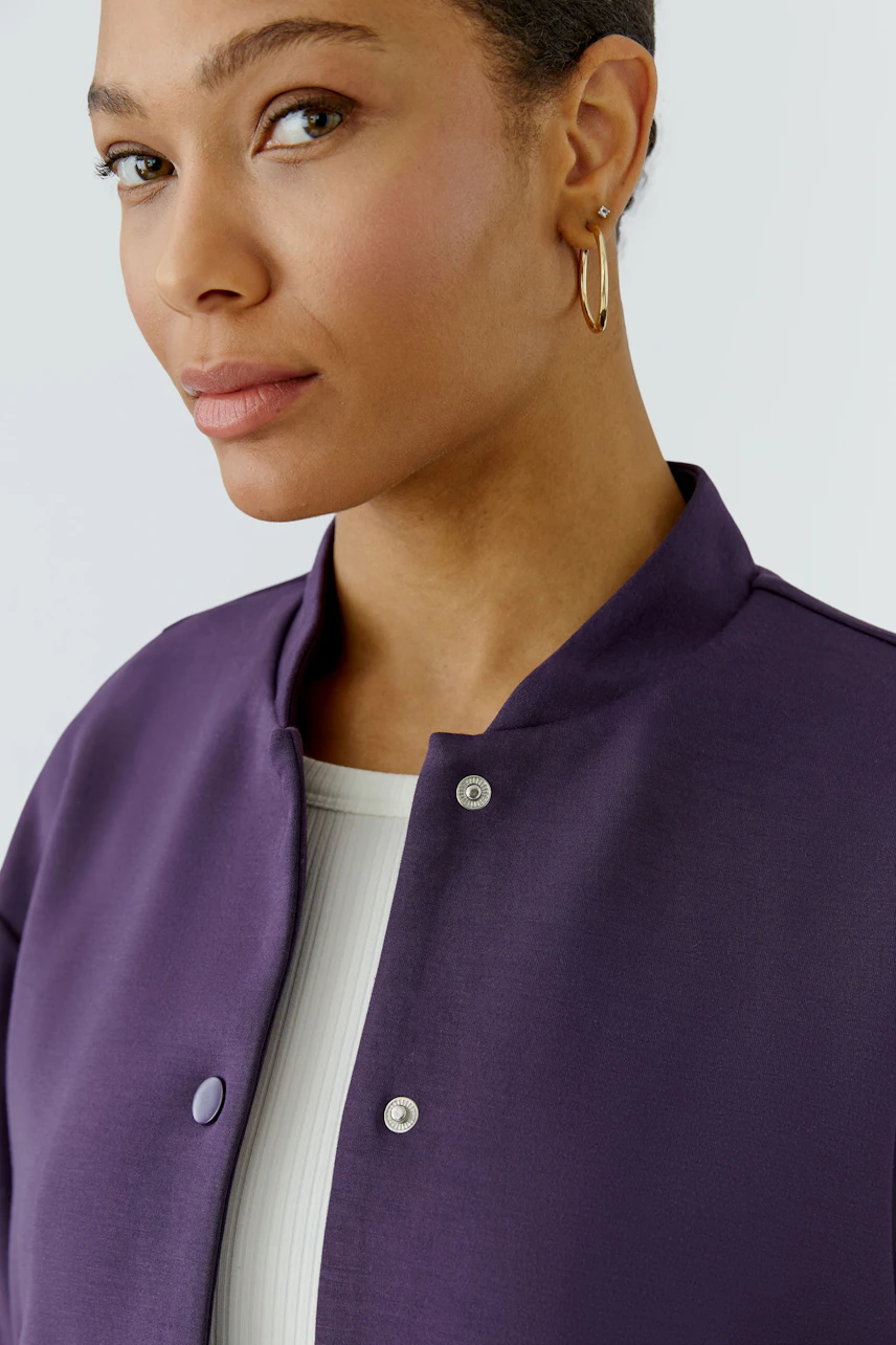 Frau im lila Blouson mit weißen Knöpfen, schlichtes Design, trägt goldene Ohrringe.