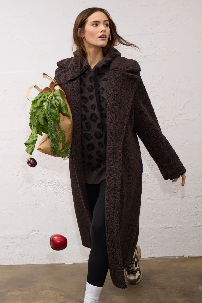 Casual winter coat: Woman in a cozy brown coat, leopard pullover, leggings, and sneakers, holding groceries.