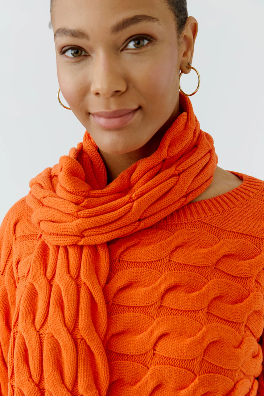 Frau mit orangefarbenem Schal und passendem Pullover, lächelt dezent. Schal mit grobem Strickmuster.