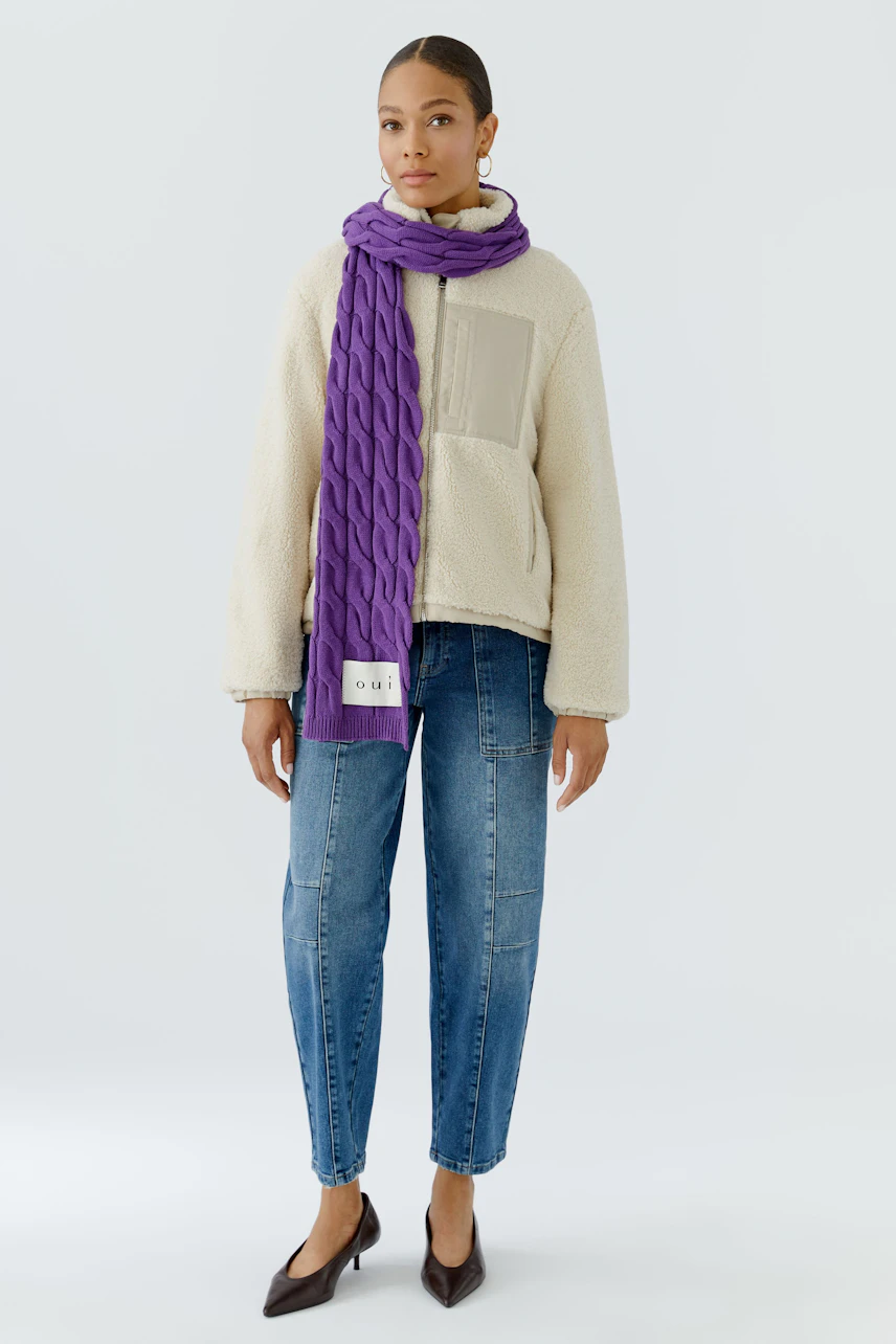 Frau mit lila Schal: Trägt beigen Pullover und Jeans, Schal locker um den Hals geschlungen.
