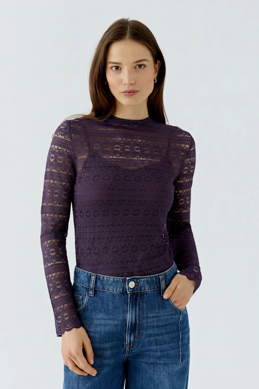 Woman in a lace long-sleeve shirt, purple, paired with blue jeans, looking confidently at the camera.
