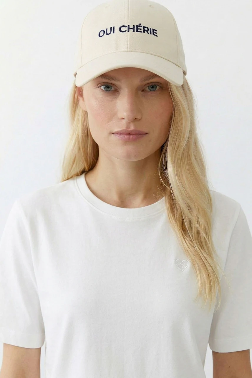 Woman in white tee wearing a beige cap with 'Oui Chérie' text, looking directly at the camera.