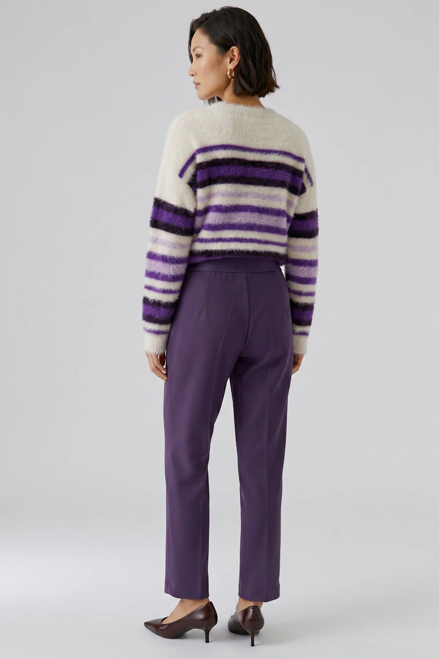 Frau in lila Culotte und gestreiftem Pullover, seitlich stehend. Lässiger Look mit Absatzschuhen.